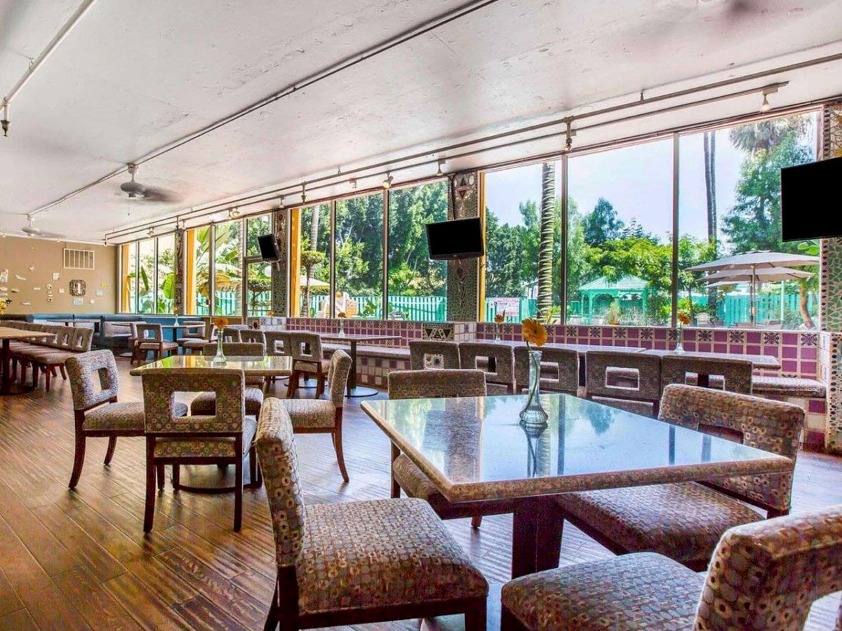 A spacious indoor dining area with multiple tables and chairs, large windows offering views of an outdoor pool area, and two wall-mounted TVs.