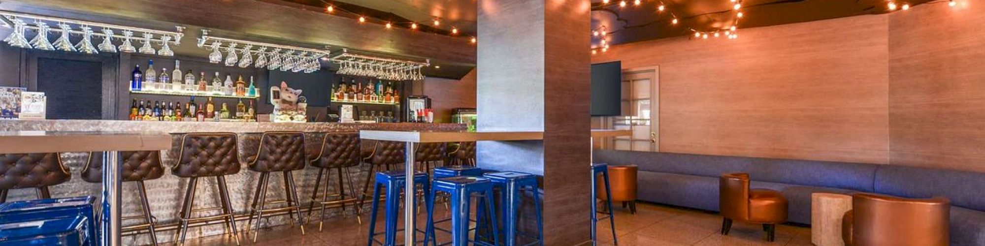 The image shows a modern bar with hanging string lights, stools at a clean counter, and seating areas with brown and blue furniture on a shiny floor.