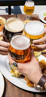 Four people toasting with beer mugs over a table filled with various dishes, including pizza, waffles, and fries, at an outdoor setting.