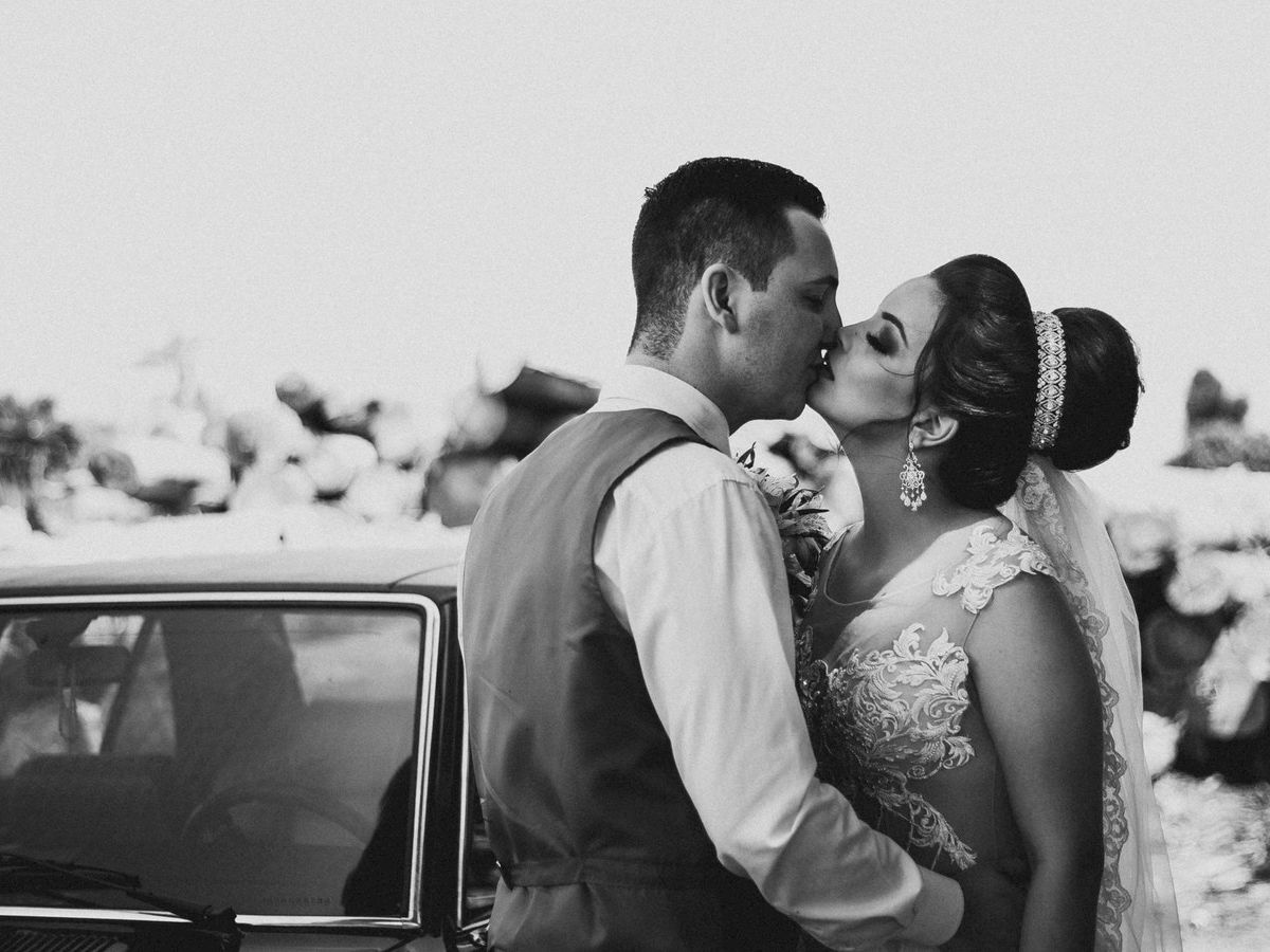 A bride and groom share a kiss beside a vintage car in a monochrome photograph, captured during their wedding celebration outdoors.