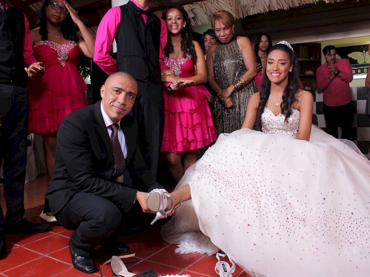 A group of people, dressed formally, gather around a young woman in a white gown, while a man kneels to help with her shoe.