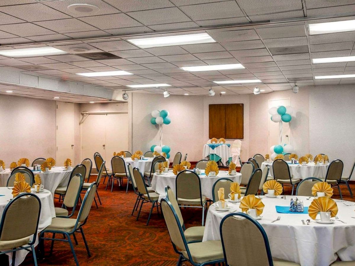 The image shows a banquet hall set up with round tables covered with white tablecloths, yellow napkins, and teal balloons, prepared for an event.