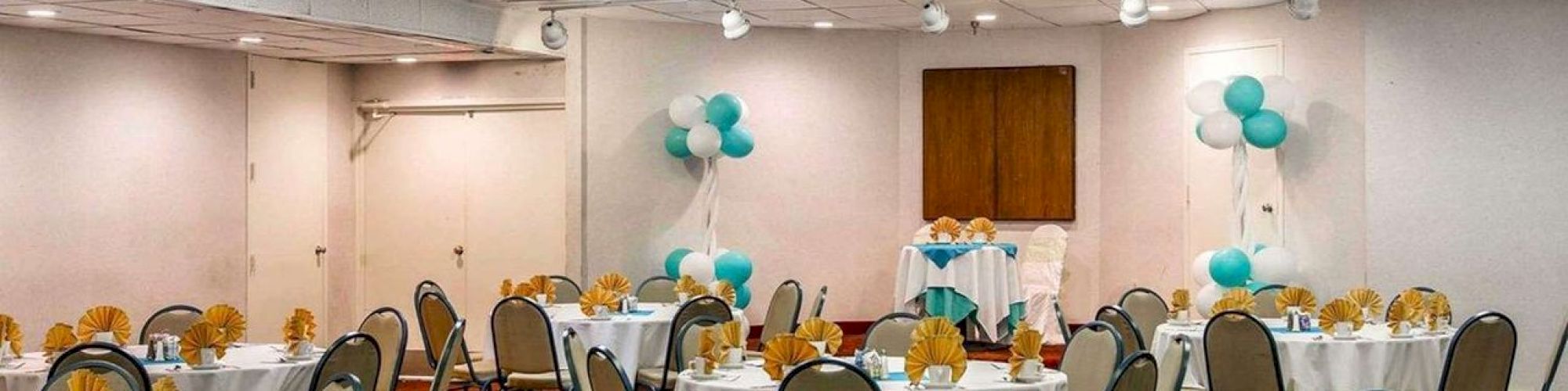 A banquet hall set for an event with round tables covered in white cloth, yellow napkins, and centerpieces, alongside balloons for decoration.