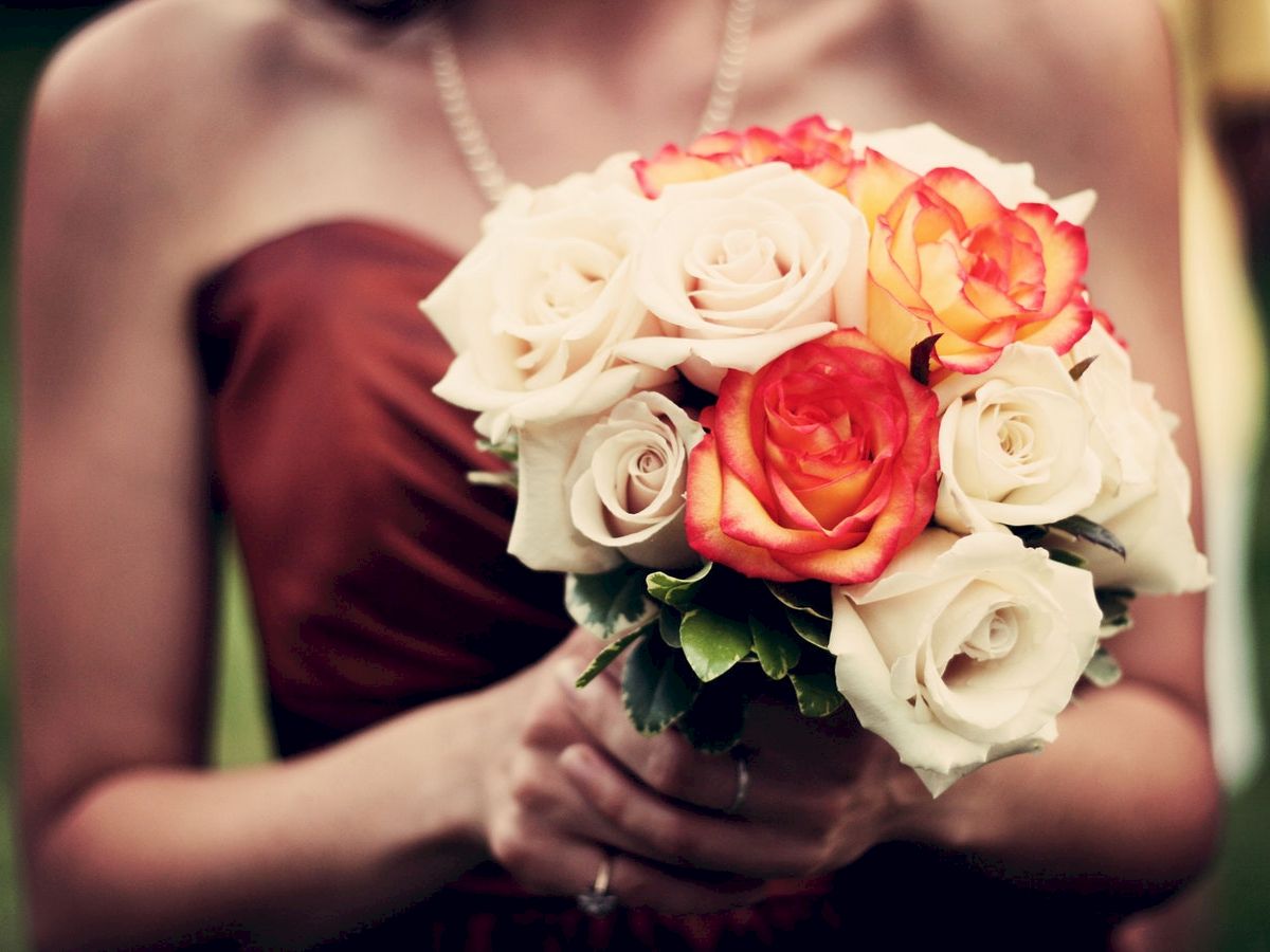 A person holding a bouquet of white and orange roses, dressed in a strapless dress and wearing a pearl necklace, with a blurred green background.