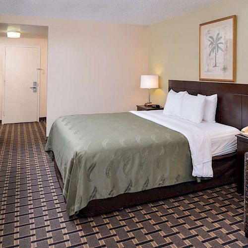 A hotel room with a queen bed, two bedside lamps, a TV on a dresser, an armchair, and a door in the background.