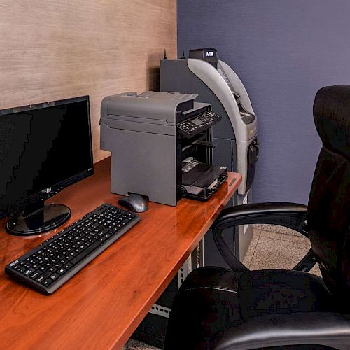 The image shows an office setup with a computer, monitor, keyboard, printer, and an ATM machine in the corner, along with a black office chair.