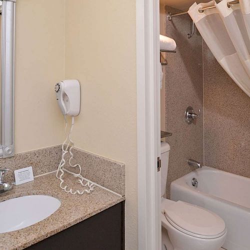 The image shows a bathroom with a sink, a wall-mounted hair dryer, a mirror, a bathtub, a shower curtain, and a white toilet.