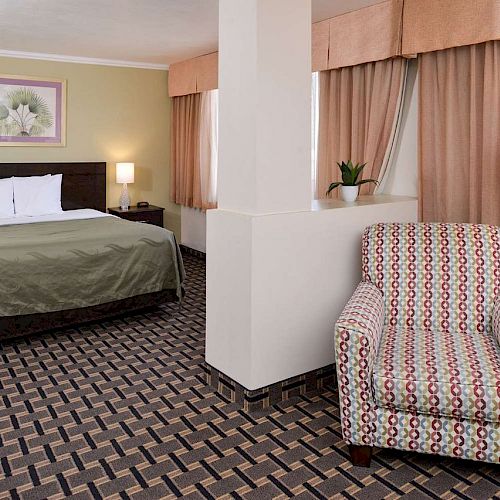 The image shows a neatly arranged hotel room with a bed, a patterned armchair, lamps, a painting above the bed, curtains, and a small plant.