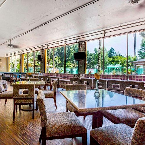 The image shows a spacious dining area with upholstered chairs and glass-top tables, large windows overlooking an outdoor pool area with umbrellas.