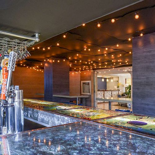 This image shows a modern bar area with various taps for beer, colorful bar countertops, and string lights hanging from the ceiling, creating a cozy atmosphere.