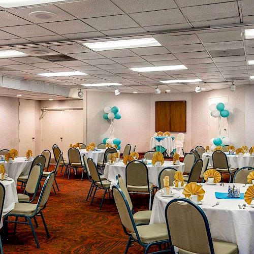 A decorated banquet hall with round tables, chairs, yellow napkins, and blue balloons, likely set up for a celebration or event.