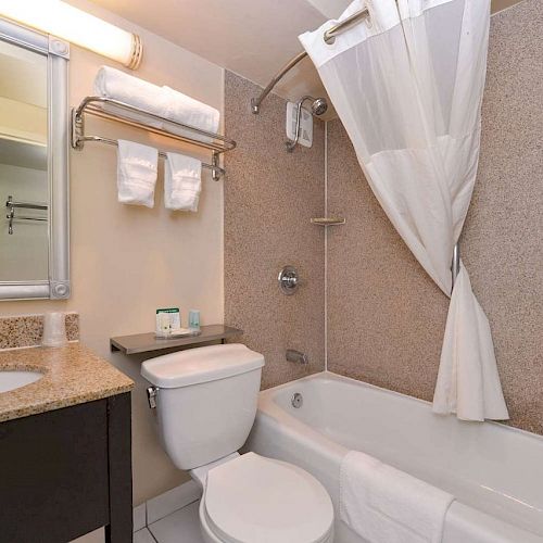 The image shows a well-lit bathroom with a sink, toilet, bathtub/shower combo, mirror, and towel rack with neatly folded towels.