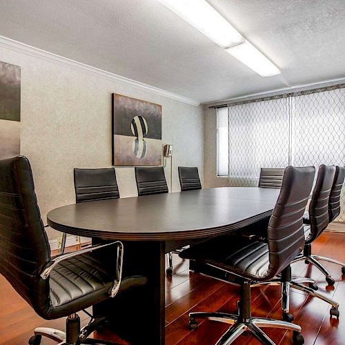 A modern conference room with a large oval table surrounded by seven black chairs, two abstract paintings, and a window with blinds ending the sentence.