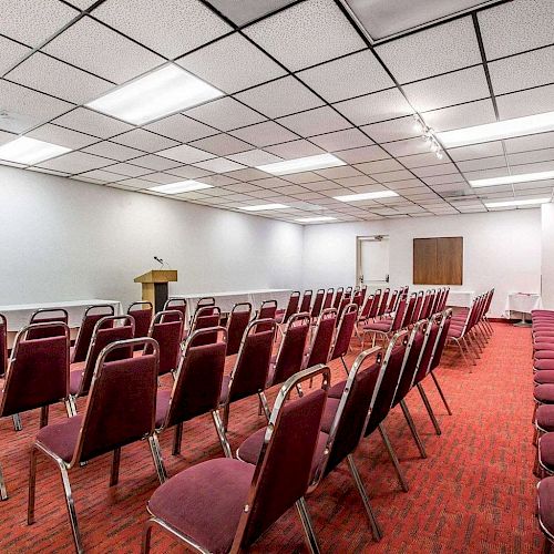 A conference room with a podium and rows of red chairs is set up for an event or meeting, featuring a whiteboard and ceiling lights.