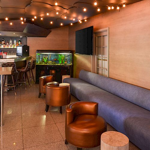 A cozy lounge with modern furniture, string lights, an aquarium, and a bar area with various seating options, including stools and armchairs.