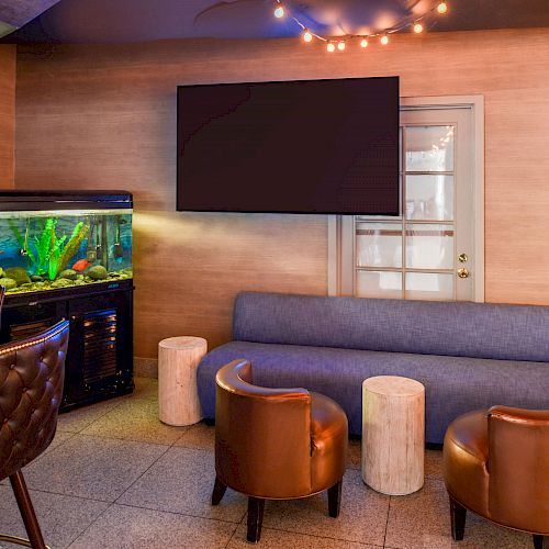 A cozy room features a large fish tank, a TV, a blue couch, brown chairs, and a bar area with stools, creating a comfortable, inviting setting.