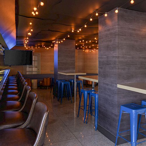 The image shows a modern bar with leather bar stools, high tables with blue stools, hanging lights, and a TV on the wall.