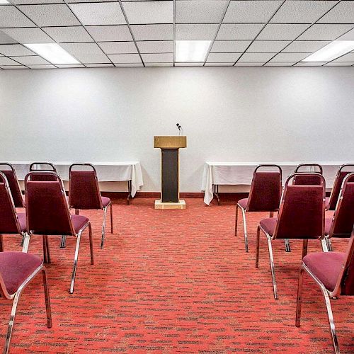 The image shows a small conference room with rows of red chairs facing a podium and microphone on a wooden stand, set against a white wall.