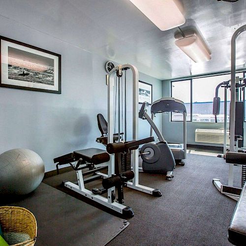 The image shows a small gym with exercise equipment such as a stationary bike, weight machine, and stability ball. There is also a window and artwork.