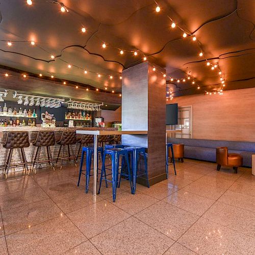 A modern bar with padded stools, sleek high chairs, string lights on the ceiling, and a spacious sitting area with cozy chairs and tables.