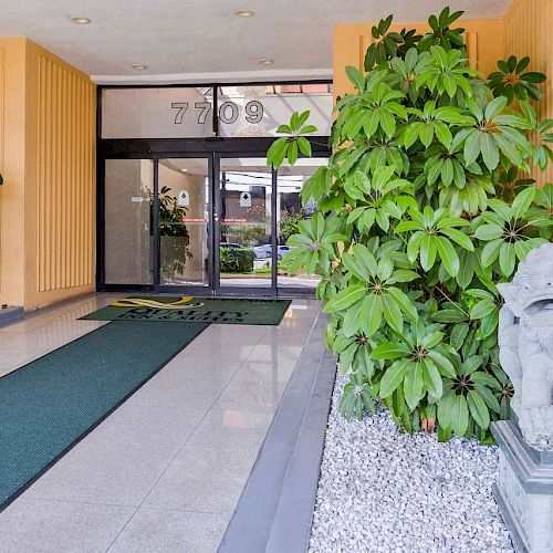 The image shows an entrance area with glass doors, greenery, a pathway with green mats, and a decorative stone lion statue to the right.