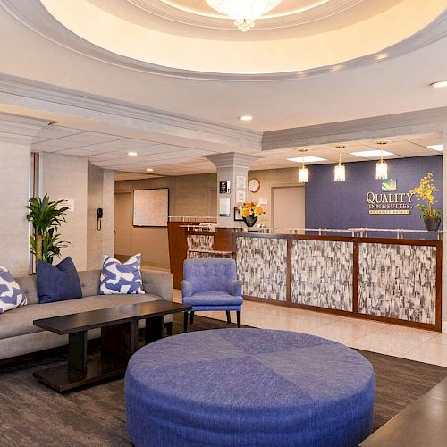 A modern hotel lobby featuring a reception desk, seating area with blue and grey furniture, decorative lighting, and plants for a welcoming atmosphere.