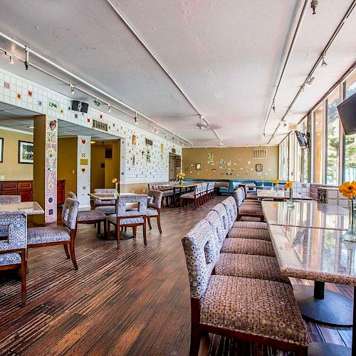 The image shows an empty dining area with patterned furniture, tables, wooden floors, large windows, and art on the walls, creating a colorful ambiance.