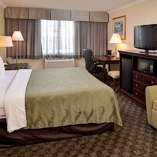 This image shows a hotel room with a bed, desk, chair, TV, and sofa. It has a warm color scheme and natural light from a window with curtains.