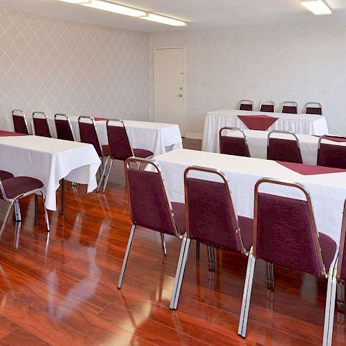 A conference room with maroon chairs, white tablecloths, and wooden flooring. Long tables are arranged facing the front.