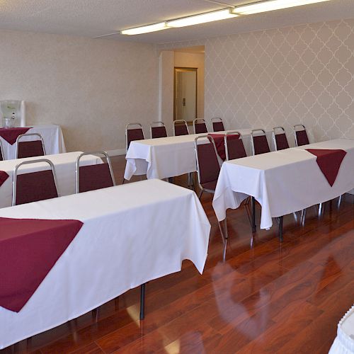 A small conference room with tables and chairs arranged in rows, white tablecloths, and burgundy napkins, with polished wood floors and patterned walls.