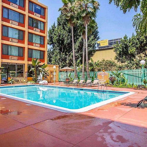 An outdoor swimming pool area with lounge chairs and tables around it, surrounded by a multi-story building and tall palm trees.