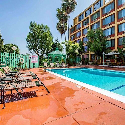 The image shows an outdoor swimming pool at a hotel with lounge chairs, greenery, and a multi-story building in the background.