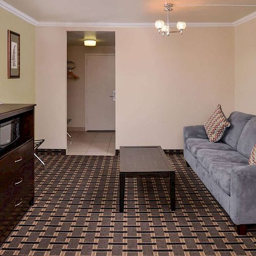 A simple living room with a sofa, coffee table, TV on a stand, and a picture on the wall. The room has patterned carpet and neutral walls.