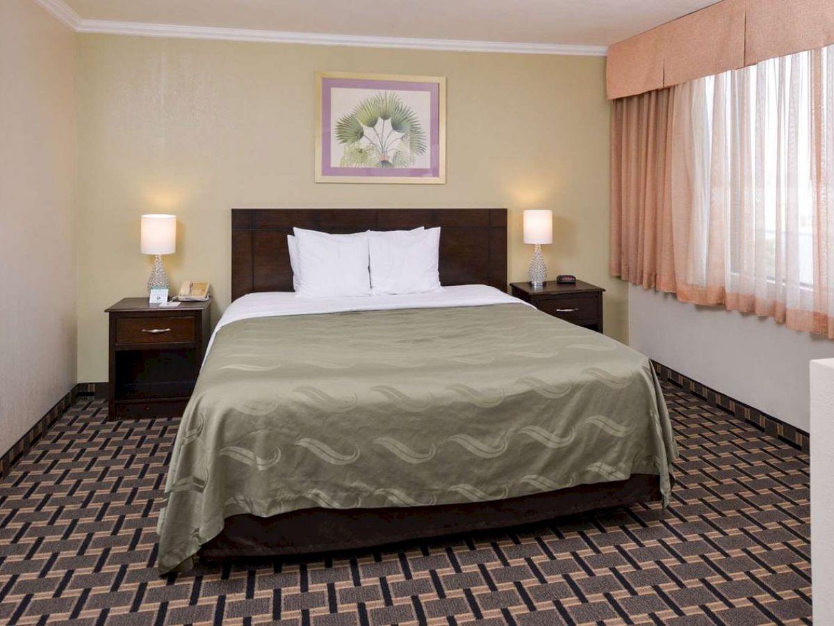 A neatly made bed in a hotel room with two bedside tables, lamps, picture on the wall, and a large window with curtains.