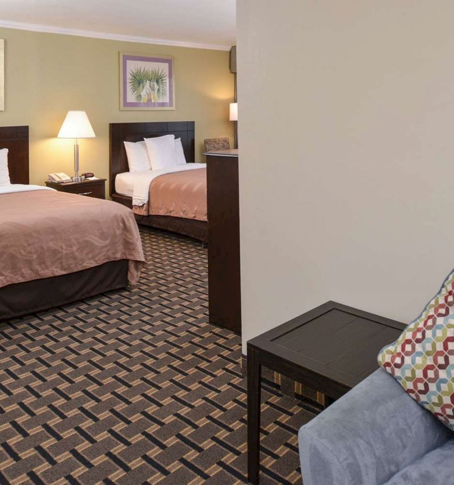 The image shows a hotel room with two double beds, a nightstand with a lamp, framed pictures, and a chair with a patterned pillow.