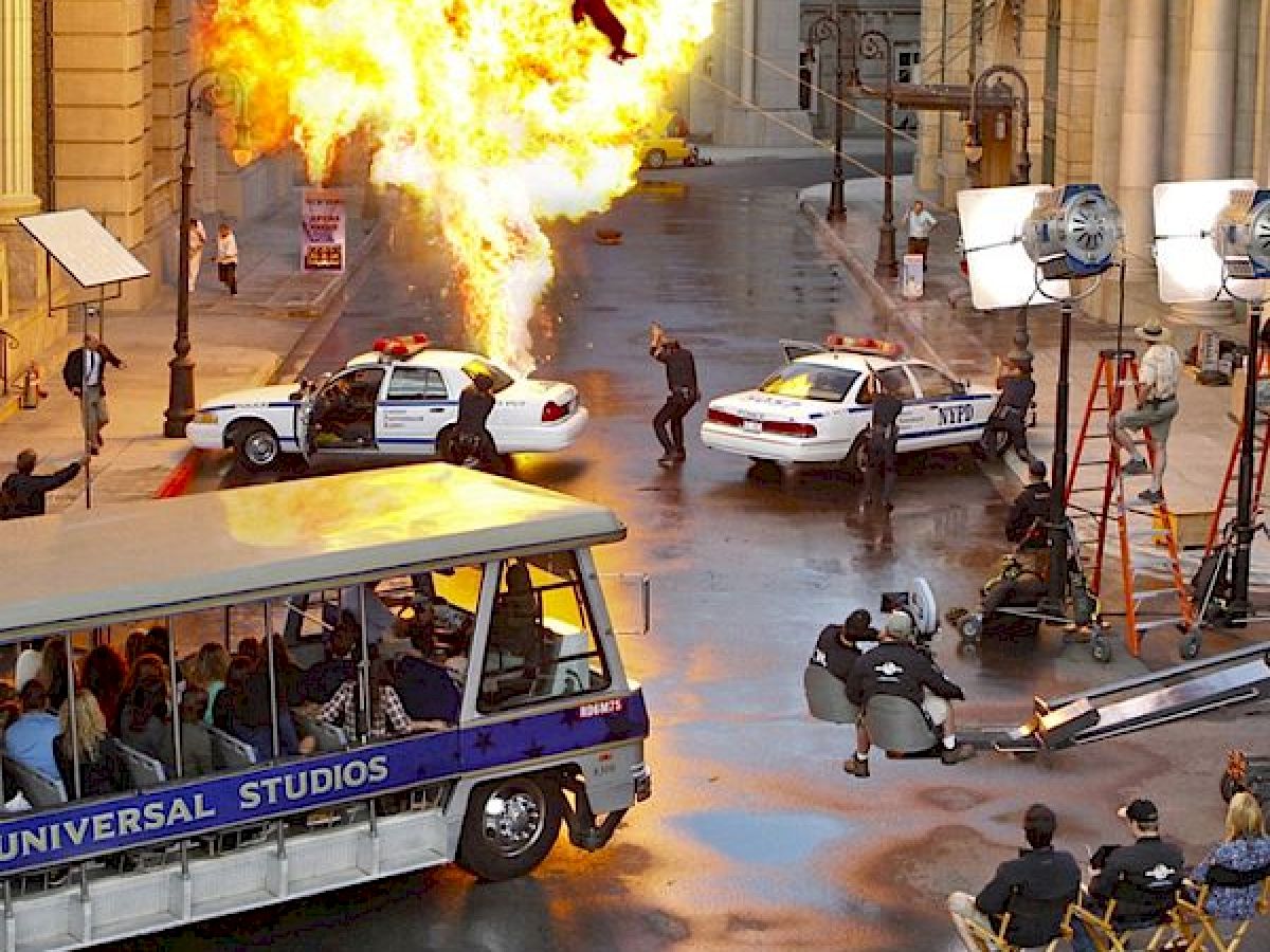 The image shows a movie set with an explosion, police cars, and crew members filming while a tour bus with people watches.