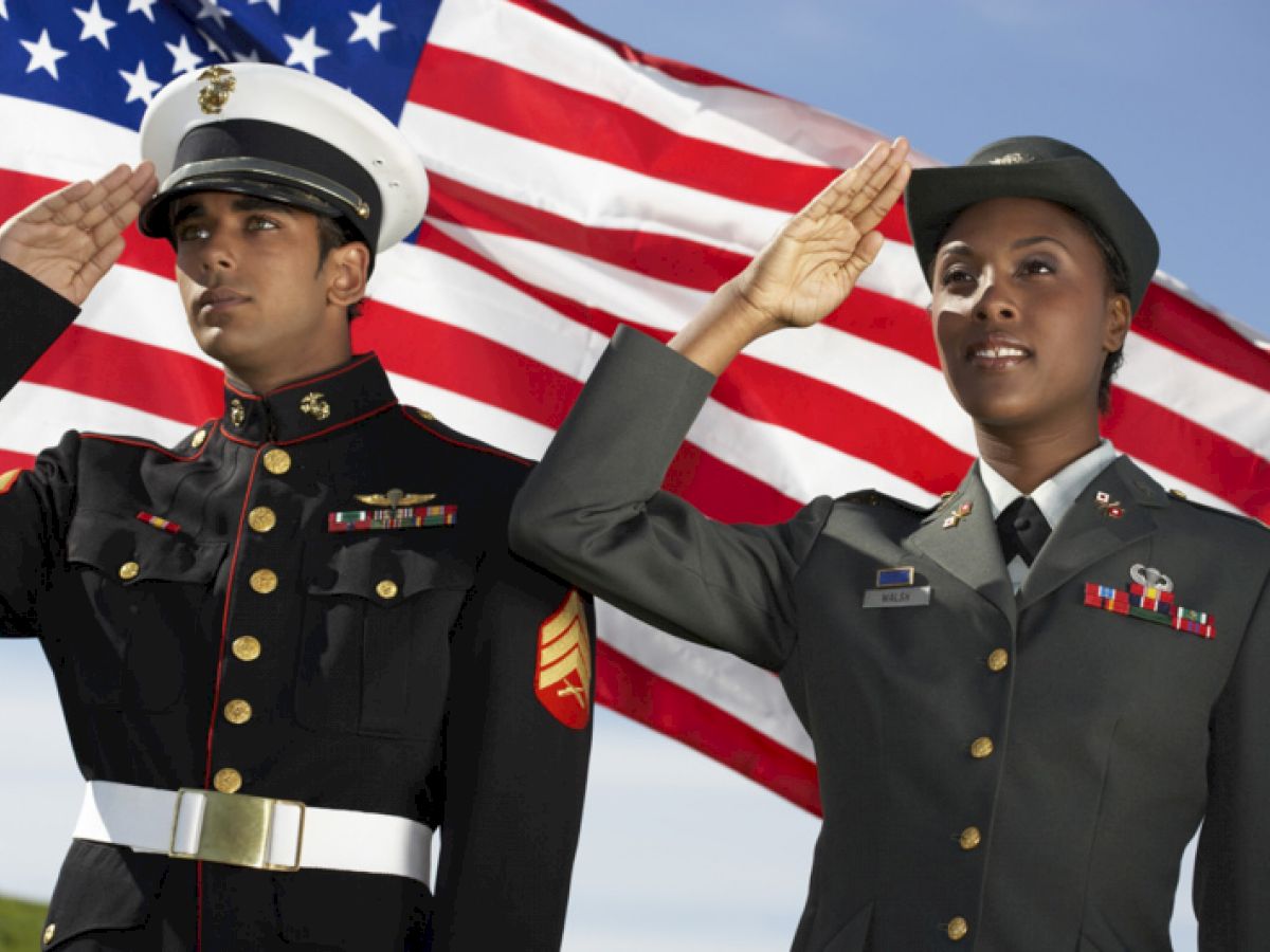 Two military personnel in uniform saluting in front of an American flag, one in a Marine uniform and the other in an Army uniform, outdoors.