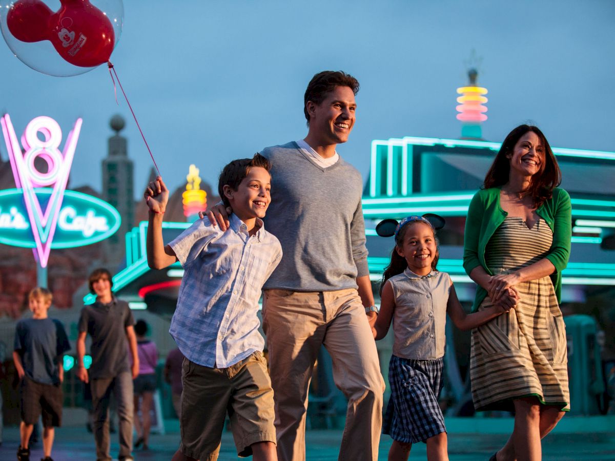 A family of four is happily walking at an amusement park at twilight, with neon signs and other visitors in the background.