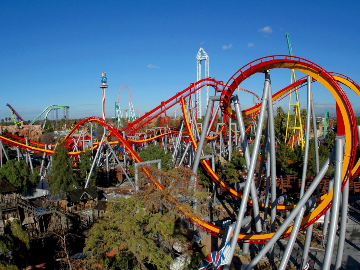 The image shows a large, intricate roller coaster with red and yellow tracks and loops, set amidst a theme park with various other attractions and rides.