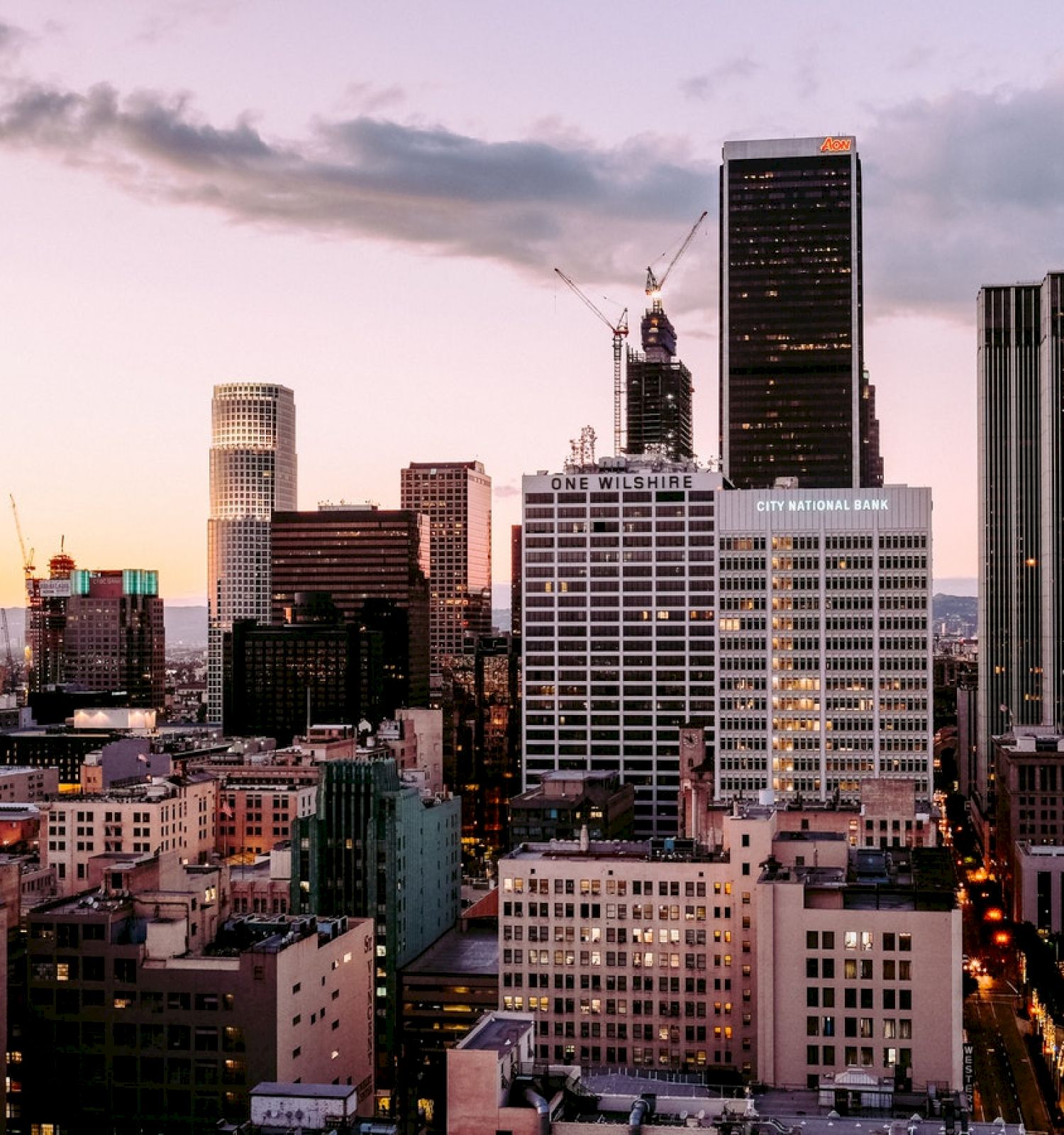 The image depicts a city skyline at dusk with various high-rise buildings illuminated against a fading sky, featuring both older and modern architecture.