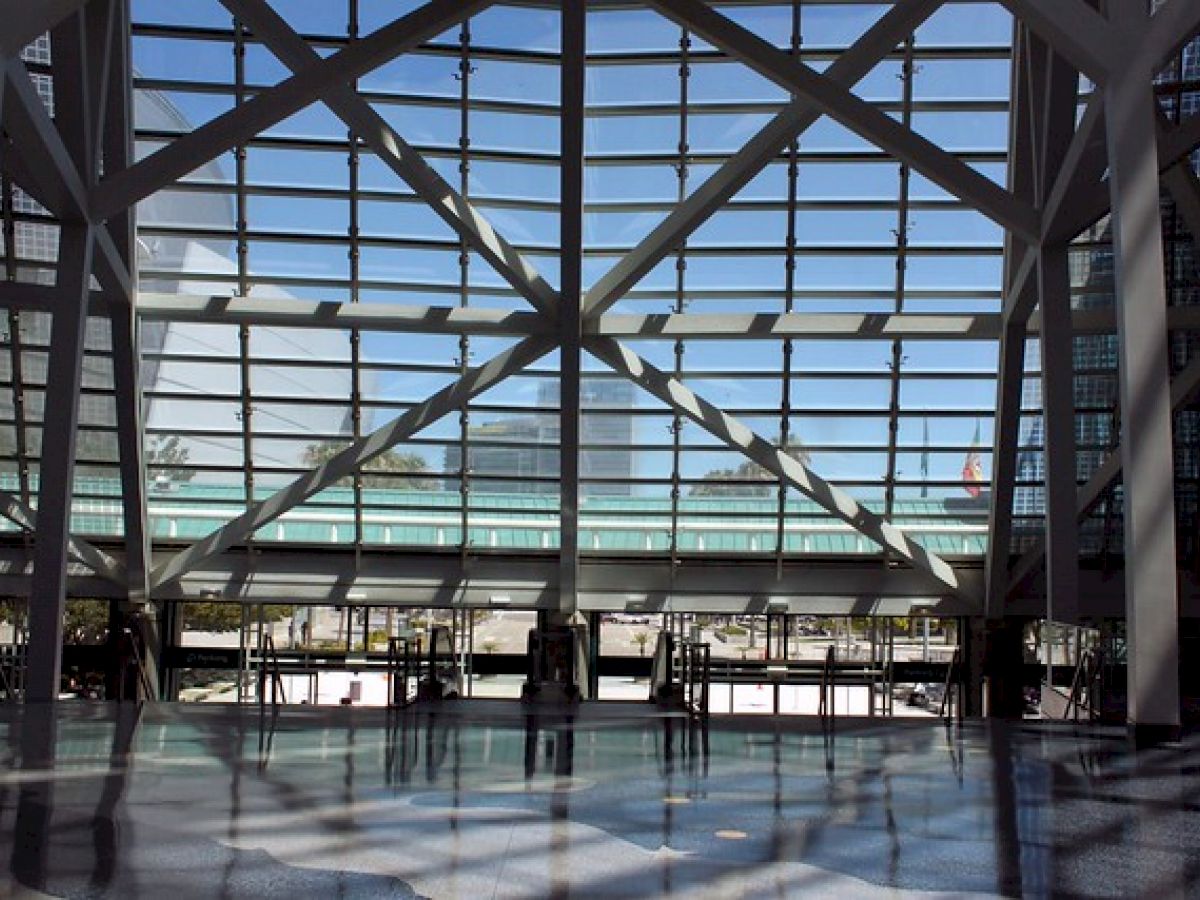 A spacious, modern indoor area with large glass windows and steel beams. People are visible outside, and light enters through the structure's grid.