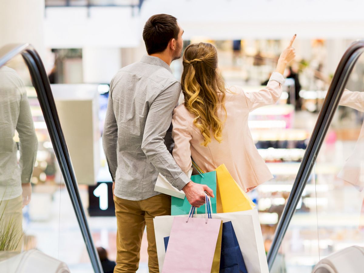 A couple is on an escalator in a shopping mall, holding multiple shopping bags, with the woman pointing towards something in the distance.