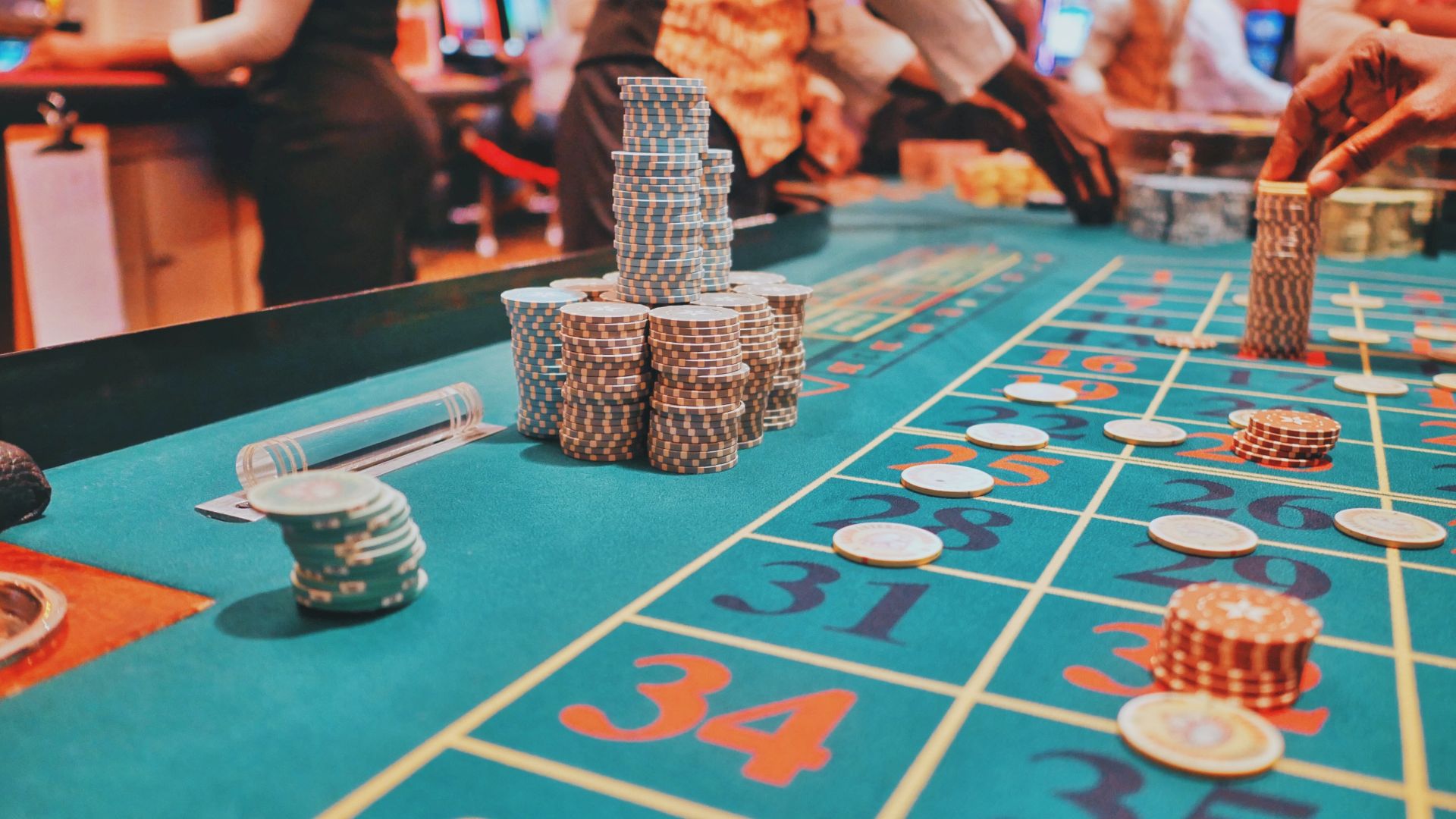 A casino roulette table with chips and player hands visible, surrounded by croupiers and other players in the background, engaging in the game.