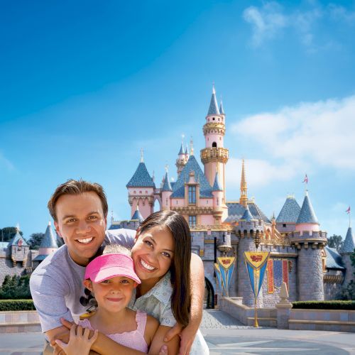 A happy family of three poses in front of a fairy tale castle at a theme park, with clear blue skies in the background.