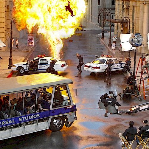 An action scene is being filmed on a movie set, with explosions, police cars, and a tour bus labeled Universal Studios, ending the sentence.