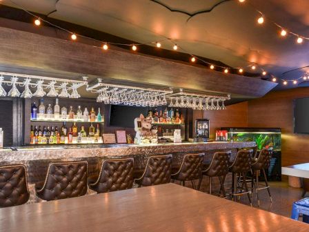 A cozy bar with a long counter, leather stools, hanging glasses, various liquor bottles, string lights, and a flat-screen TV in the background.