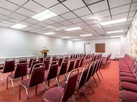 This image shows an empty conference room with rows of red chairs facing a podium, under fluorescent ceiling lights.