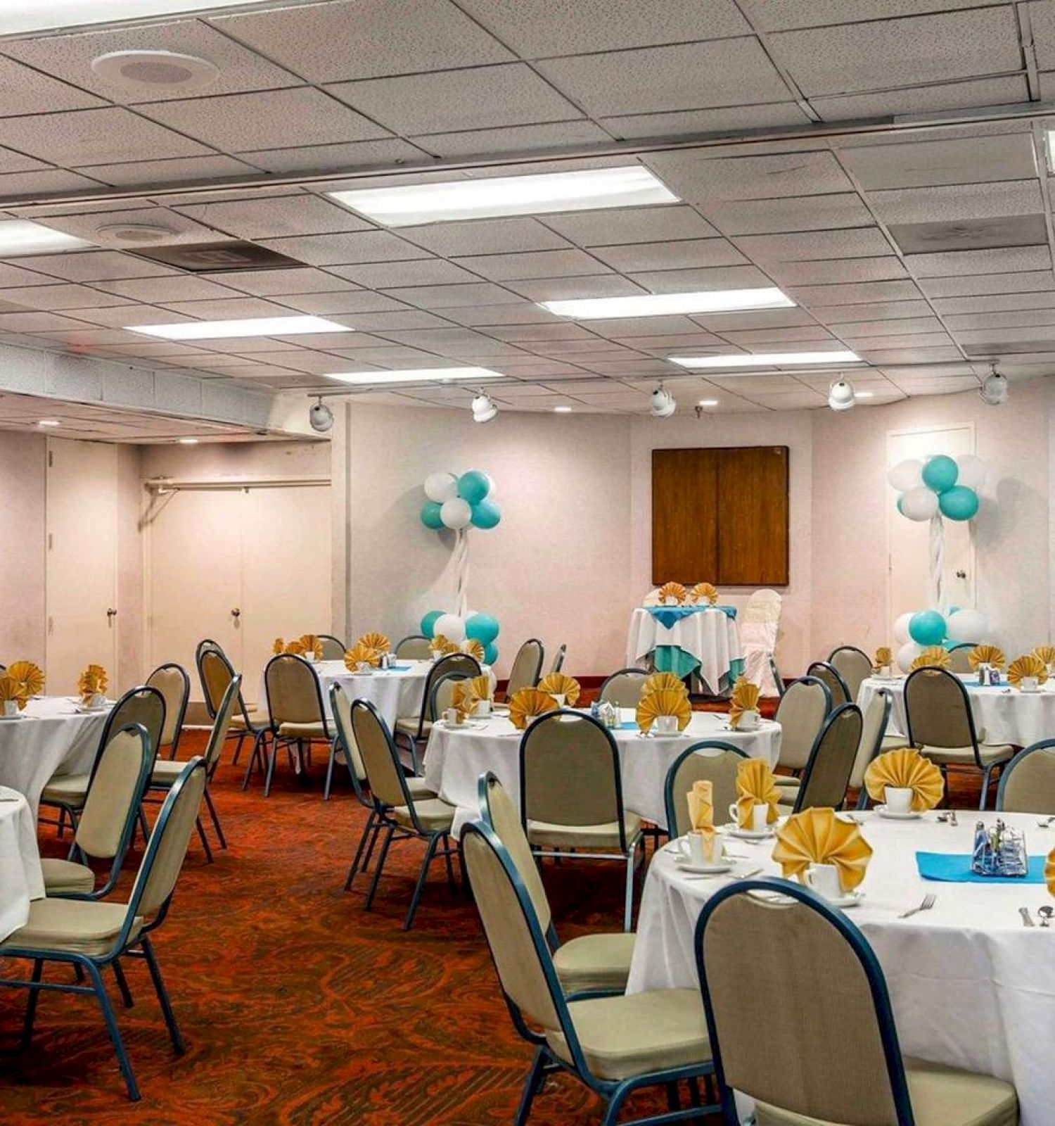 The image shows a banquet hall set up for an event with round tables and chairs, decorated with yellow napkins and blue balloons.