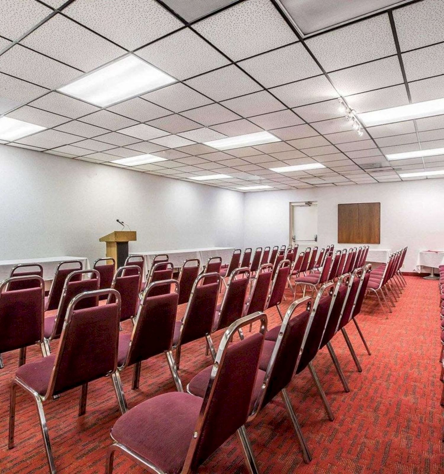 The image depicts a conference room set up with rows of red chairs facing a podium, with white walls and a red carpeted floor.