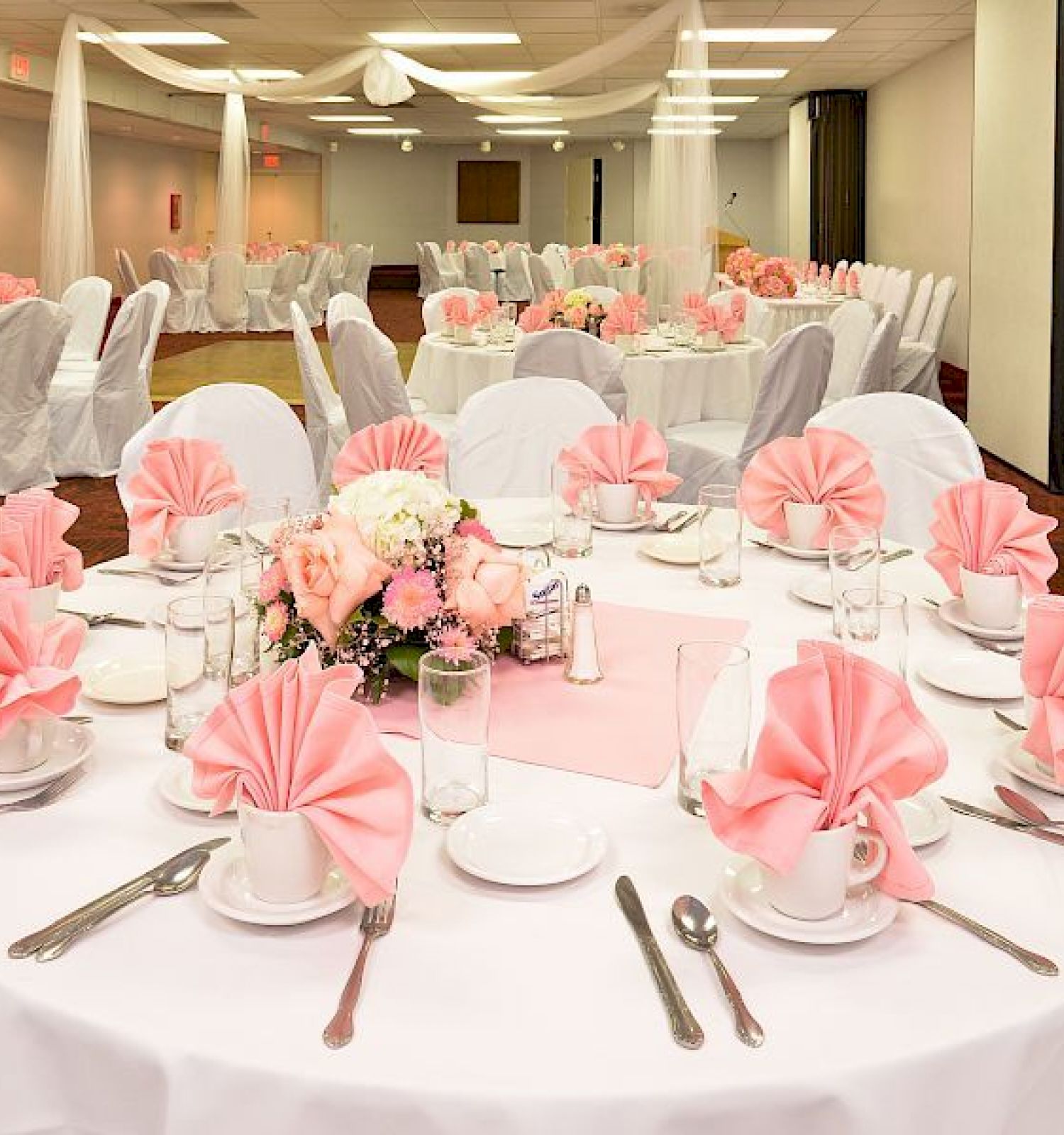 A decorated banquet hall with white tablecloths, pink napkins, floral centerpieces, and multiple tables set for an event or celebration.